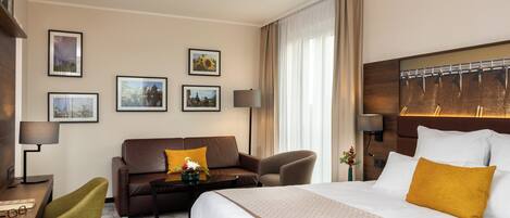 A hotel room with a large bed, a sofa, a desk with a lamp, and framed pictures on the wall.