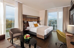 A hotel room with a bed, a desk, a chair, a small table with a plant, and a view of the city.