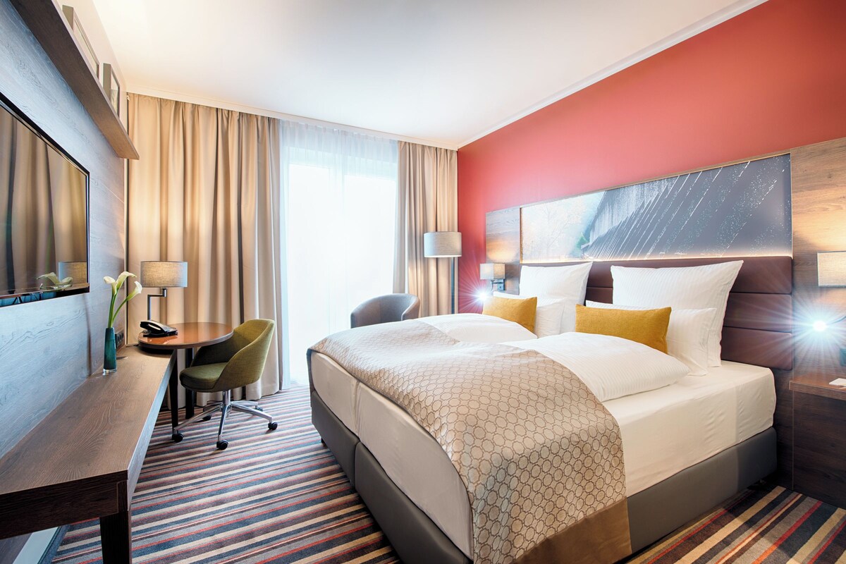 Quarto de hotel com uma cama grande, uma escrivaninha, uma cadeira, um abajur e uma janela ampla com cortinas.