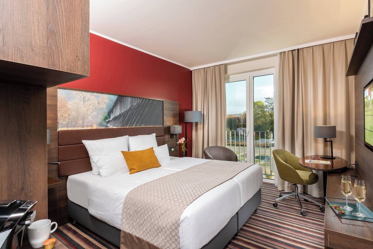 A hotel room with a large bed, a desk with two chairs, a TV, and a balcony with curtains.
