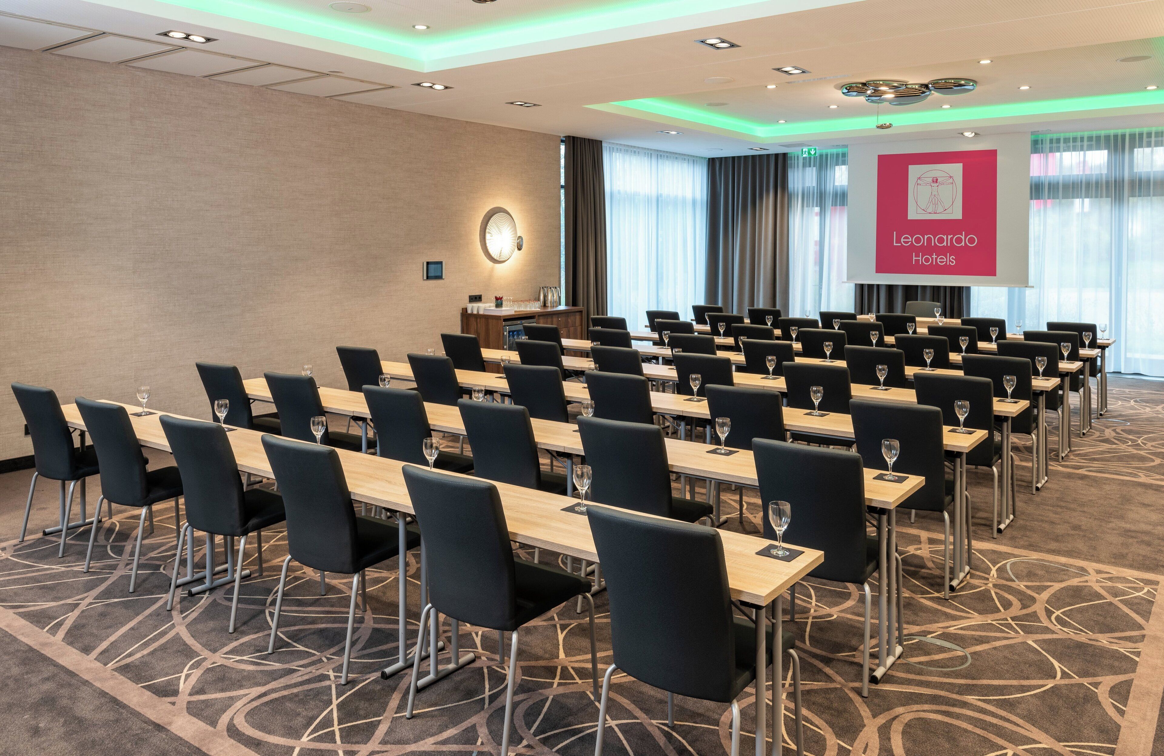 A conference room with rows of tables and chairs, a podium, and a projector screen displaying the logo of Leonardo Hotels.