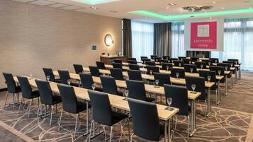 A conference room with rows of tables and chairs, a podium, and a projector screen displaying the logo of Leonardo Hotels.