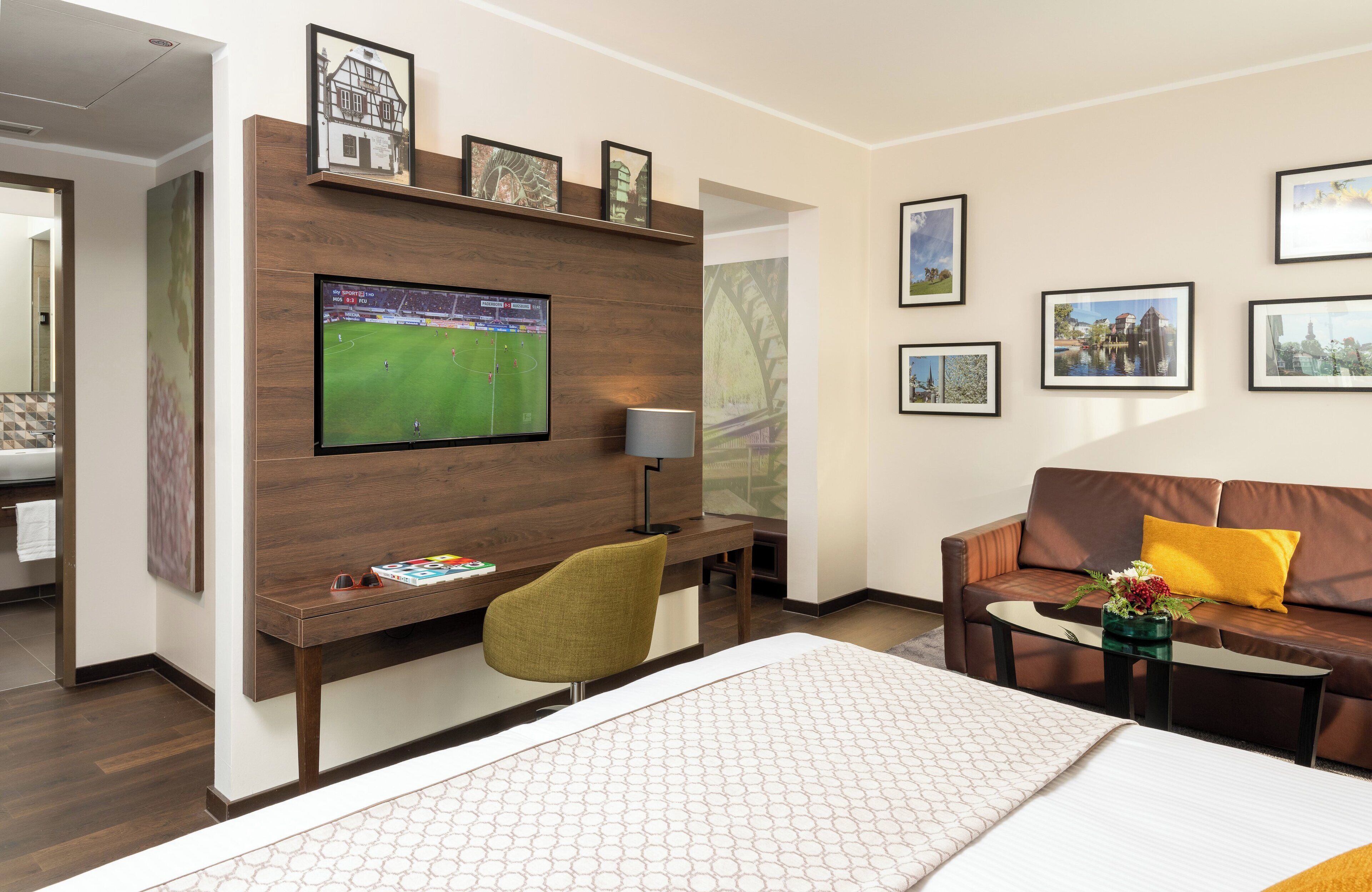 A hotel room with a large bed, a desk with a computer, a TV mounted on the wall, a sofa, and framed pictures on the wall.