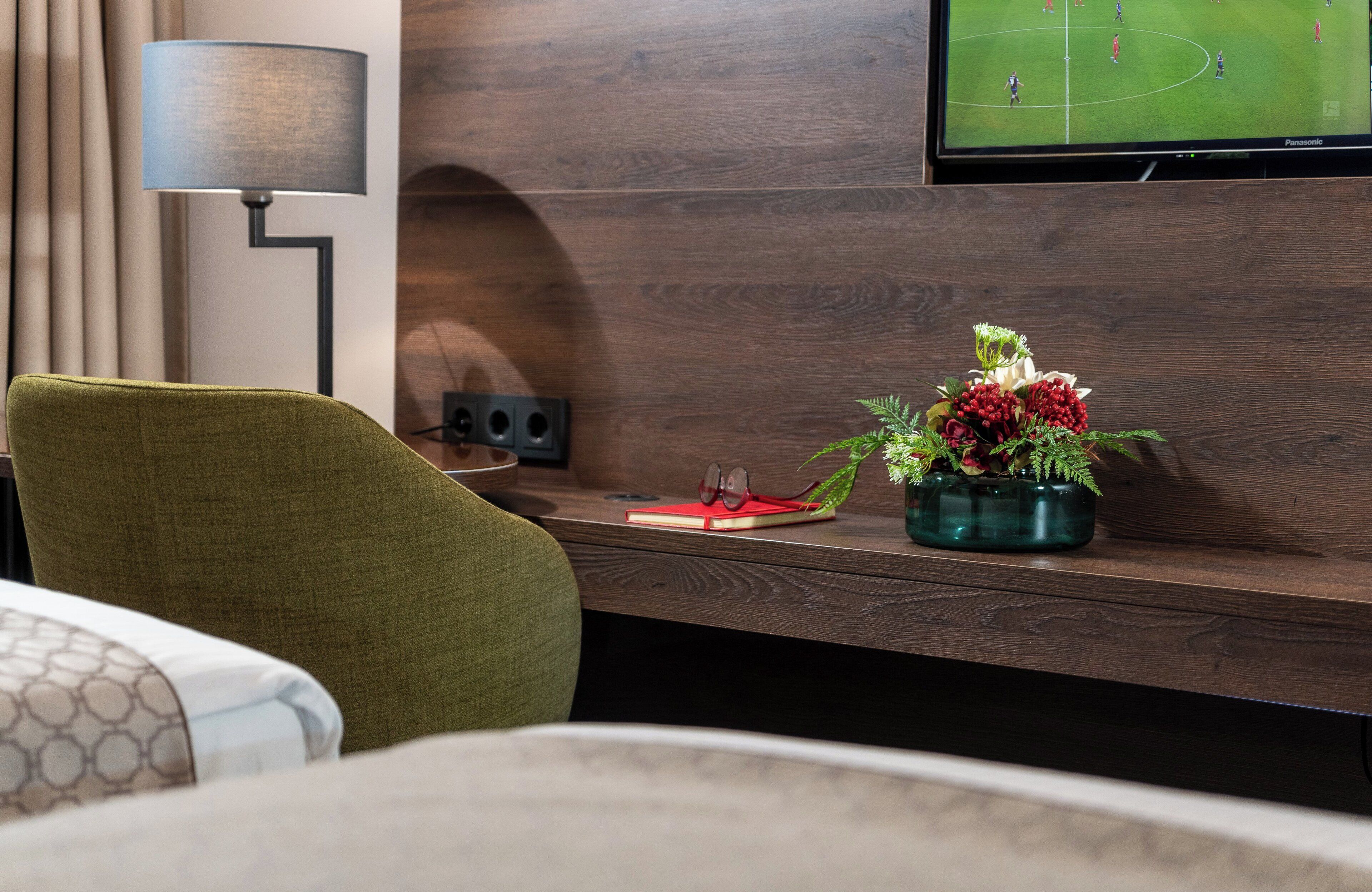 A hotel room with a wooden desk, a lamp, a TV displaying a sports game, a green chair, a red notebook, and a vase with flowers.