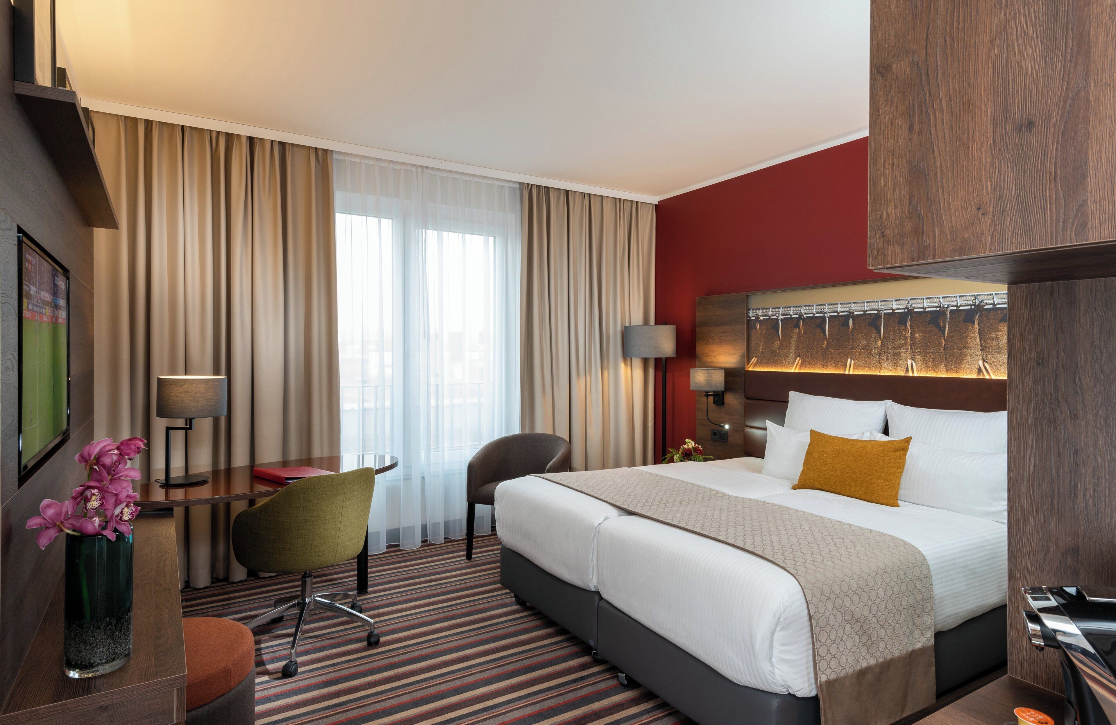 Ein Hotelzimmer mit einem Bett, einem Schreibtisch, einem Stuhl, einer Lampe und einem großen Fenster mit Vorhängen.