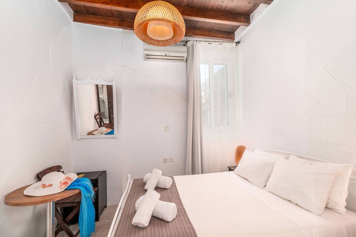 A bedroom with a bed, a bedside table, a chair, a hanging light fixture, and a window with curtains.