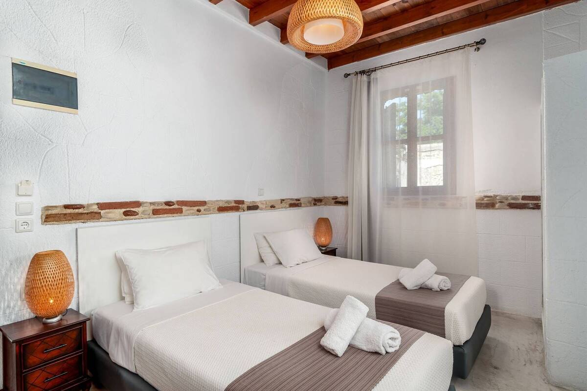 A hotel room with two beds, a wooden headboard, a brick accent wall, a window with sheer curtains, and a wall-mounted light fixture.