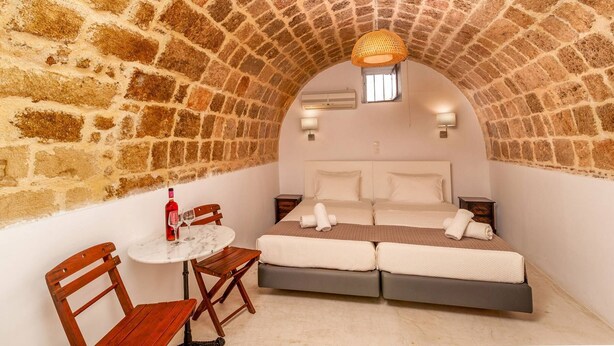 A cozy room with a stone arch ceiling, a bed with white linens, a small round table with a bottle and glasses, and a wooden chair. - Auberge 32 | Old Town (Rhodes)