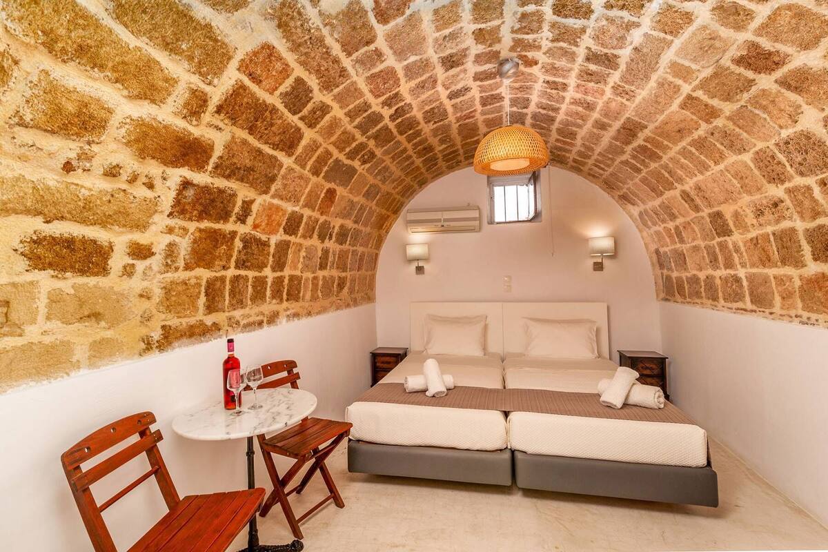 A cozy room with a stone arch ceiling, a bed with white linens, a small round table with a bottle and glasses, and a wooden chair.