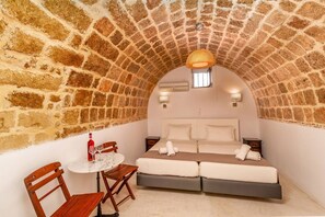 A cozy room with a stone arch ceiling, a bed with white linens, a small round table with a bottle and glasses, and a wooden chair. - Auberge 32 | Old Town (Rhodes)
