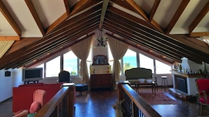 An attic room with exposed wooden beams, a fireplace, a television, and a view of the sea through large windows. - Margarita Apartments (Preveza)