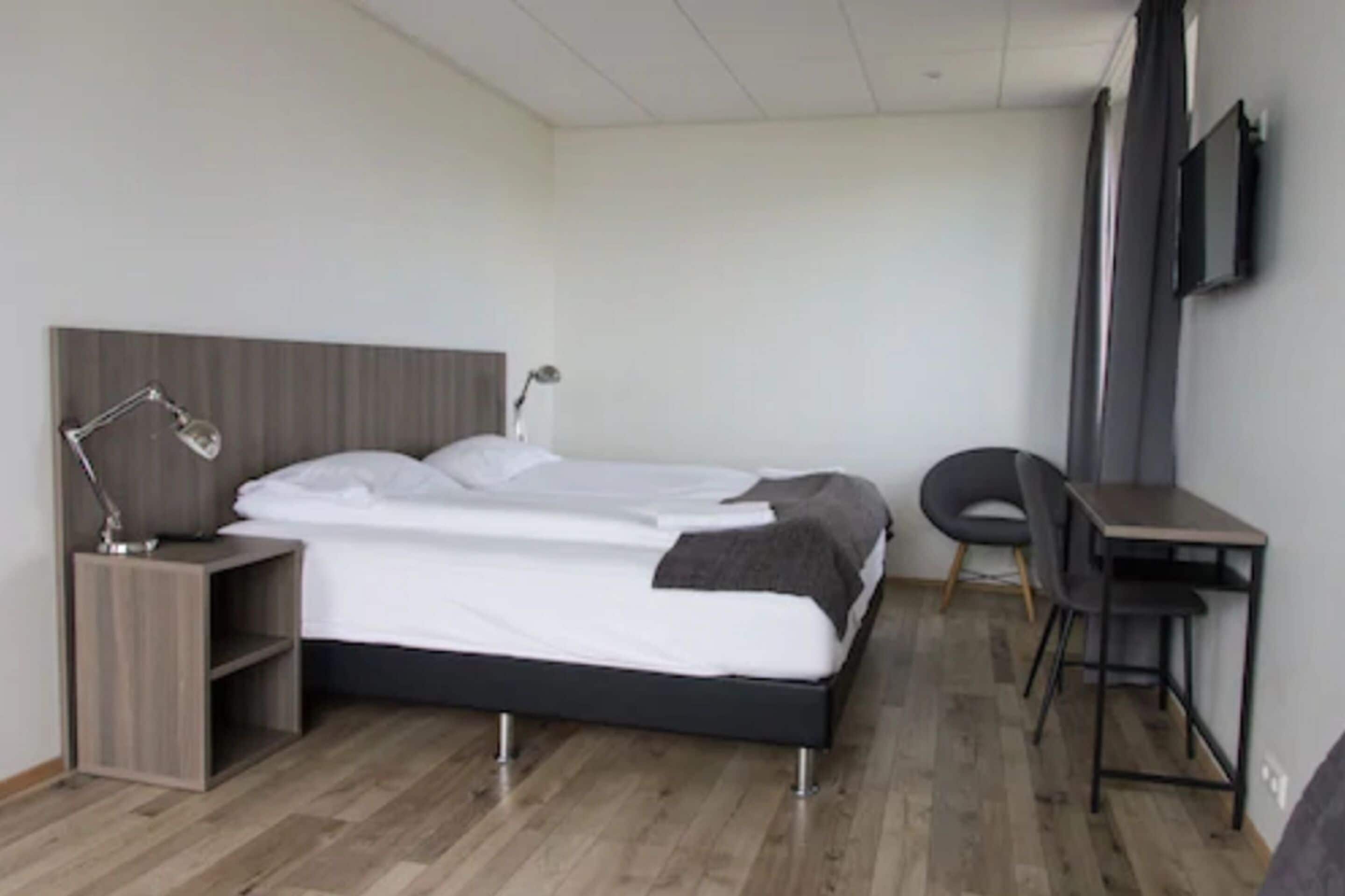 A modern hotel room with a bed, bedside table, lamp, chair, desk, and TV.