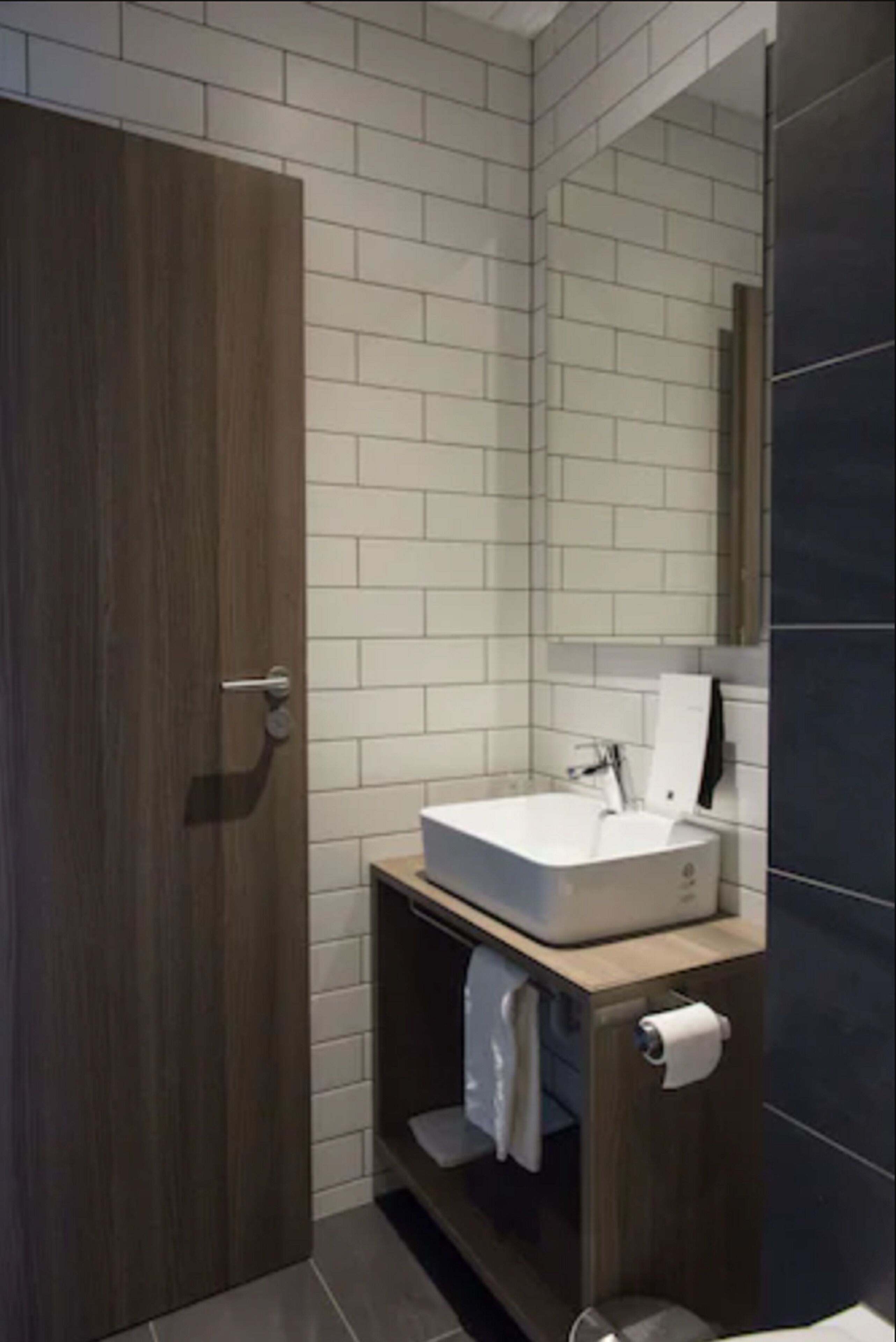 A modern bathroom with a wooden door, white tiled walls, a white sink, and a mirror.