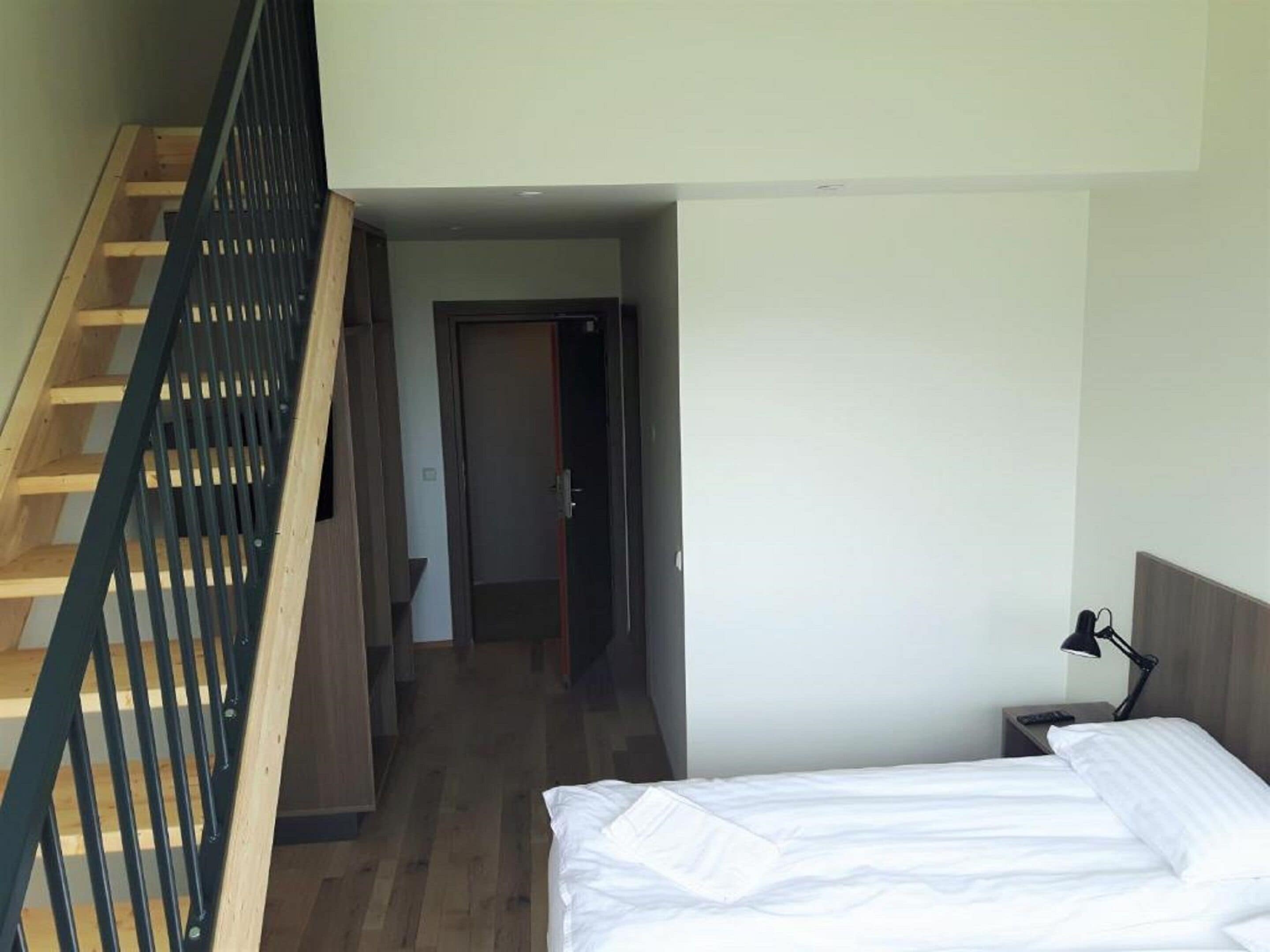 Un dormitorio con una escalera de madera, una cama con ropa de cama blanca, una mesita de noche con una lámpara y una puerta que da a otra habitación.
