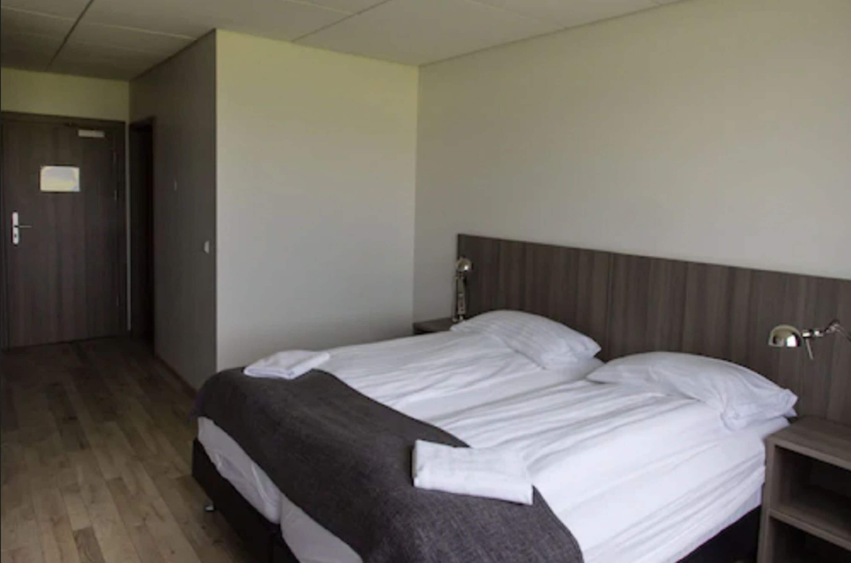 A hotel room with a double bed, wooden flooring, a wardrobe, and a bedside table with a lamp.