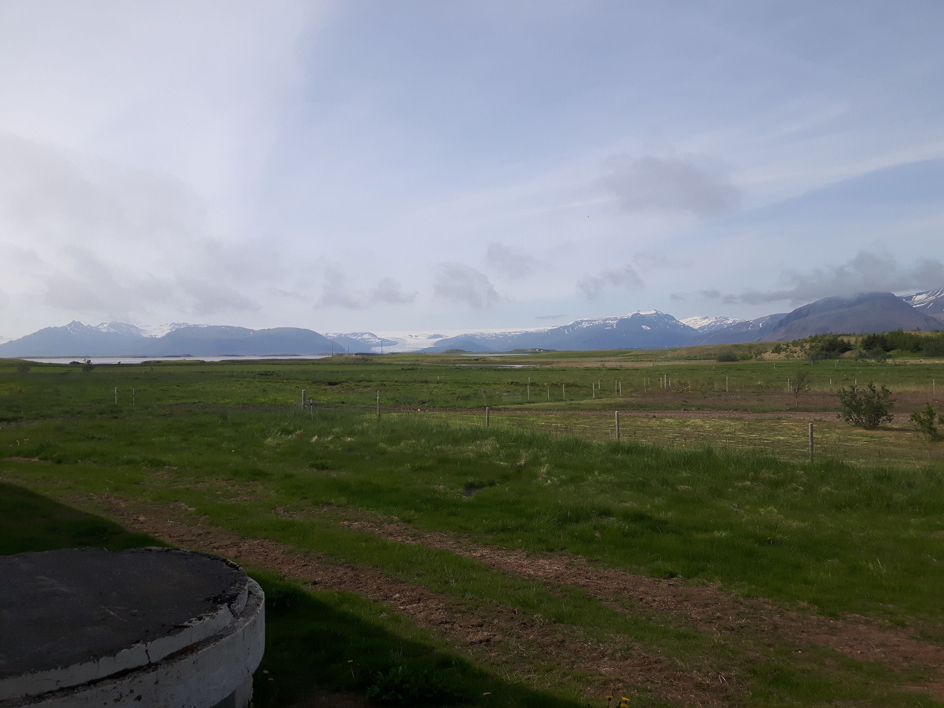 A rural landscape with a grassy field, a fence, and distant mountains with snow caps.