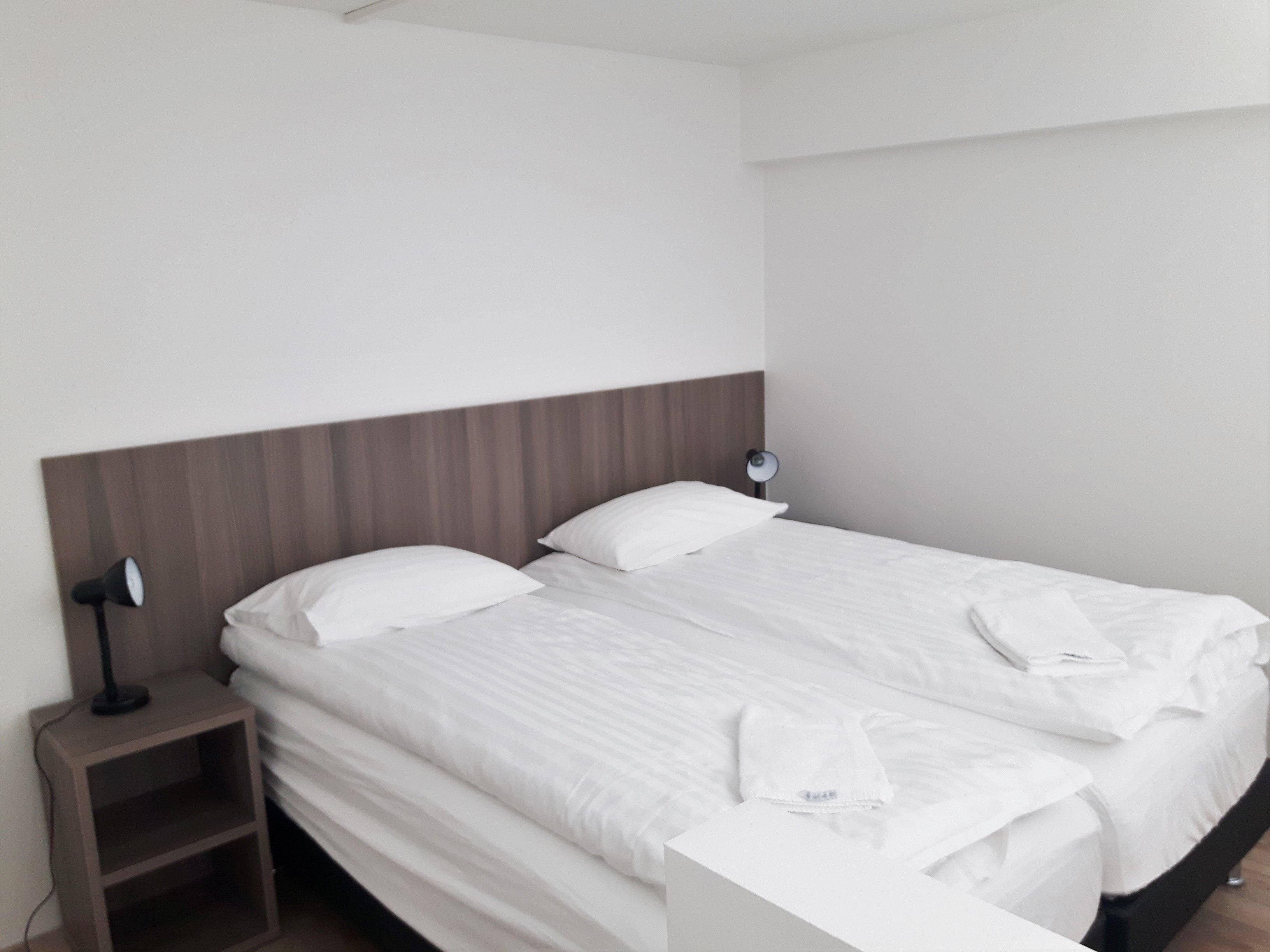 Una cama bien hecha con sábanas blancas, una lámpara de noche y una mesita de madera.