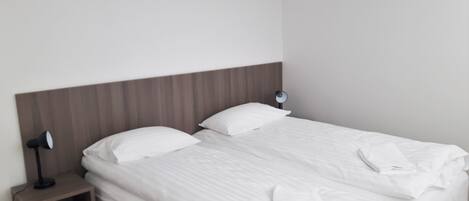 Una cama bien hecha con sábanas blancas, una lámpara de noche y una mesita de madera.