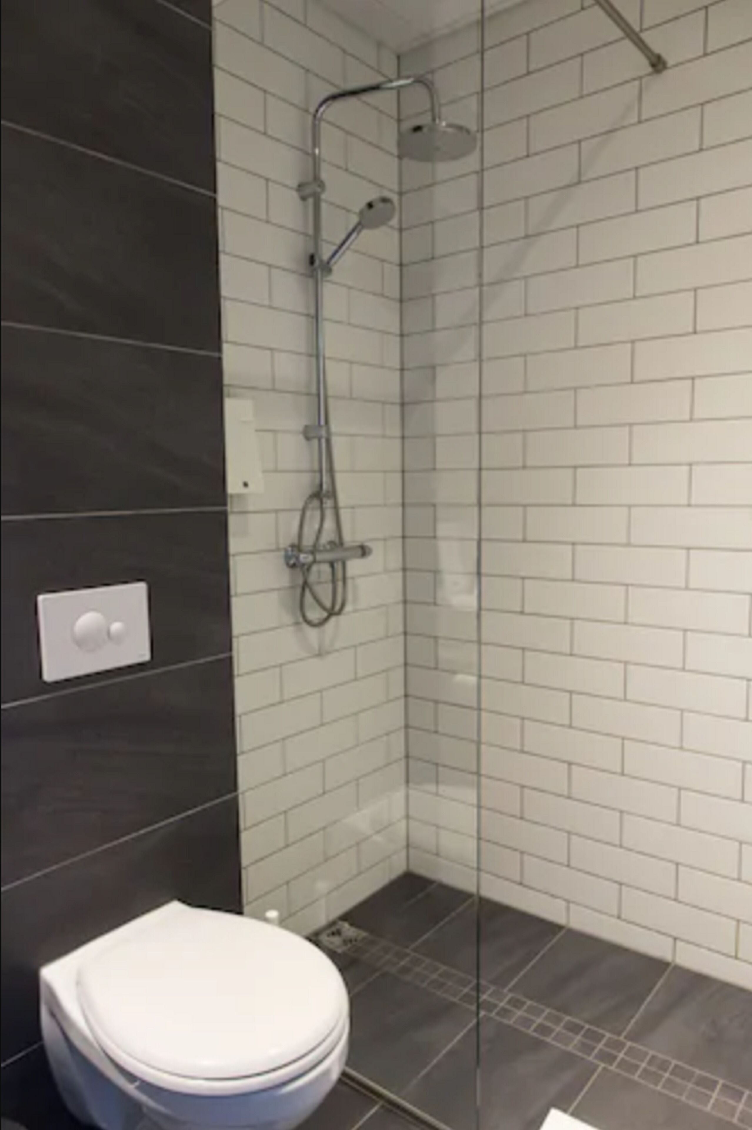 Une salle de bain moderne avec une douche à l’italienne en verre, des murs recouverts de carreaux blancs et un sol foncé.