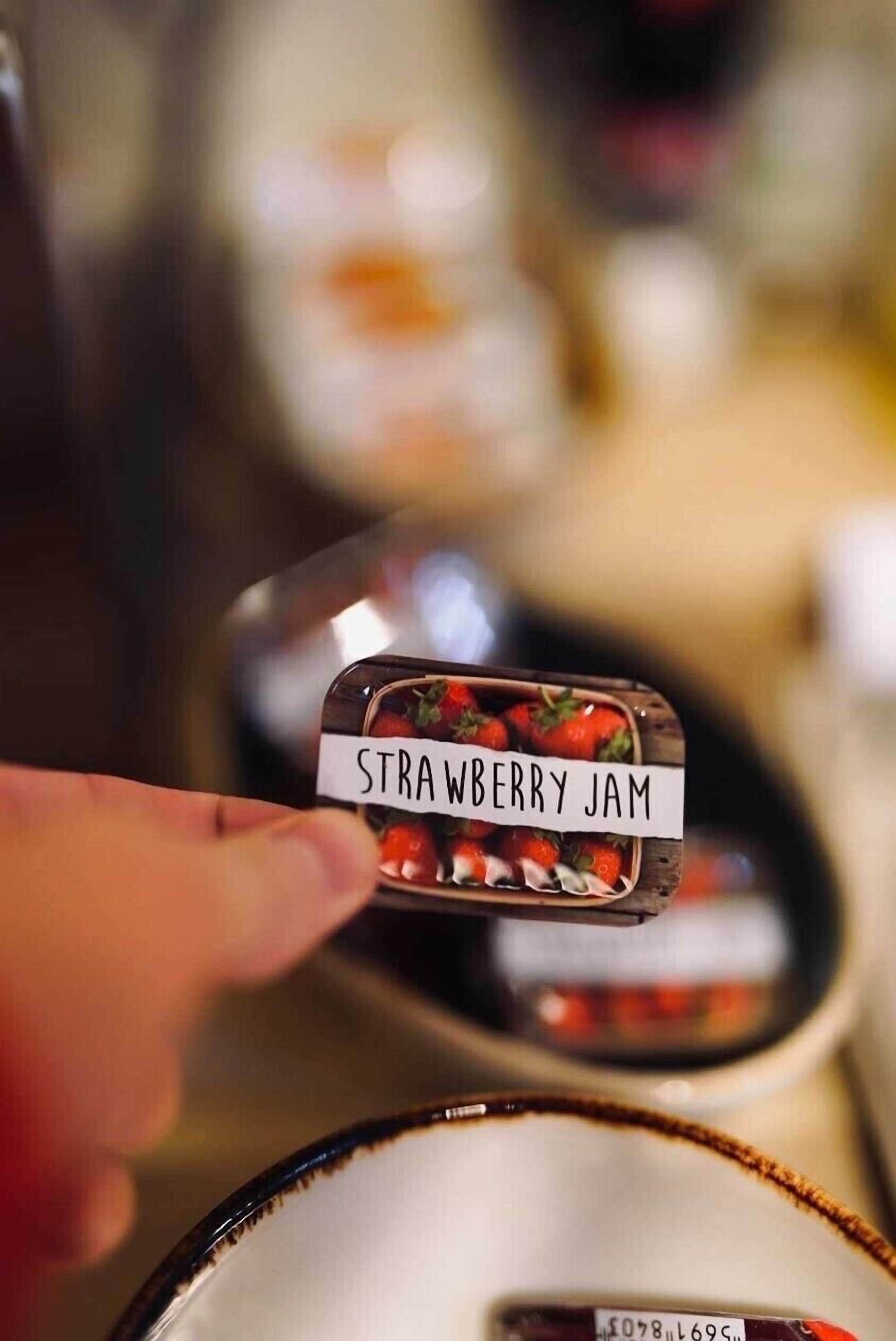 A hand holding a container labeled "STRAWBERRY JAM" with strawberries visible inside.