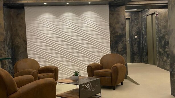 A modern lobby with two brown armchairs, a glass-top coffee table, and a textured wall.