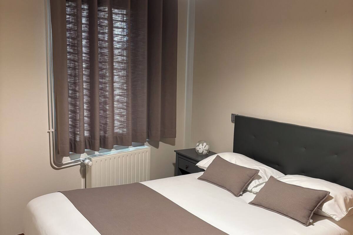 Un lit bien fait, recouvert d’une couvre-lit blanche et marron, avec une tête de lit sombre et une table de chevet sur laquelle se trouve une horloge.