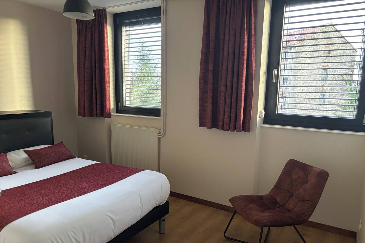 Une chambre d’hôtel avec un lit, une chaise et deux fenêtres avec des rideaux.