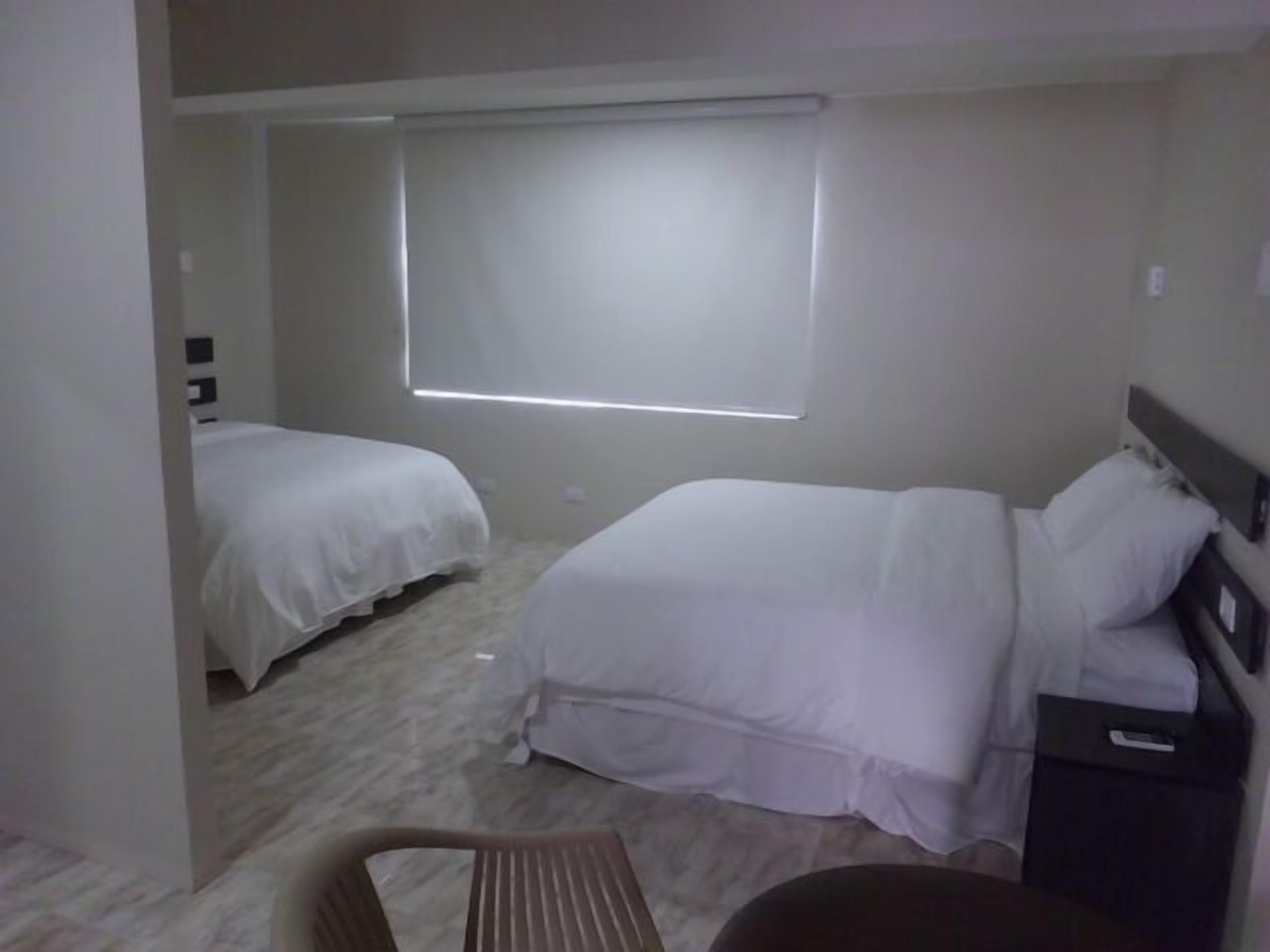 A hotel room with two beds, a window with blinds, a chair, and a nightstand with a remote control.