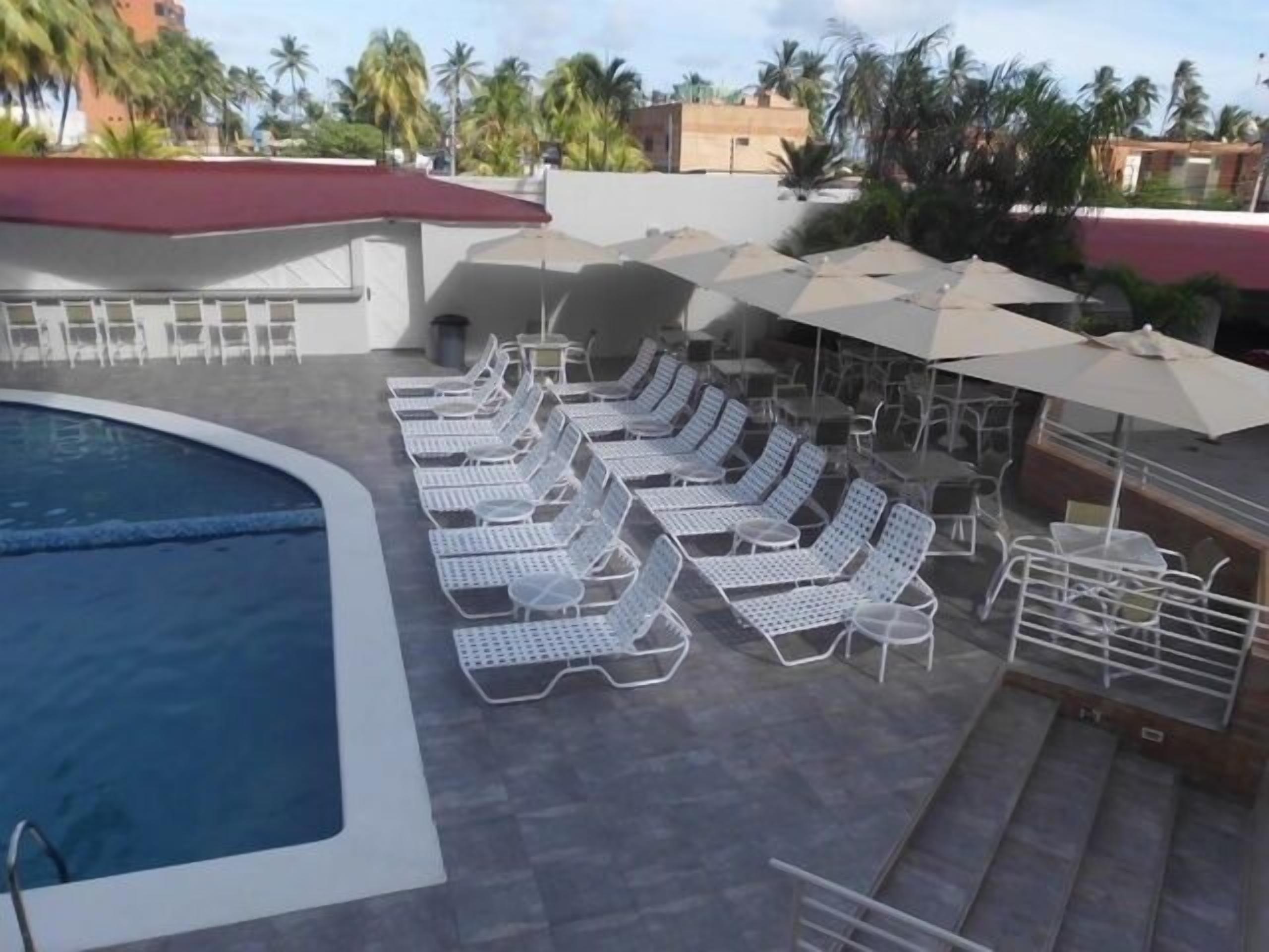 A pool area with sun loungers and umbrellas, surrounded by palm trees and buildings.