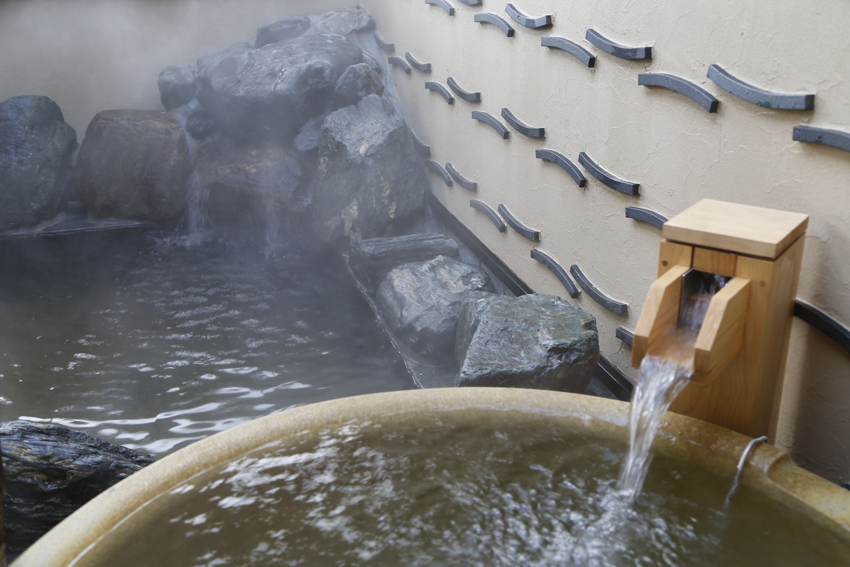 Un baño de aguas termales con agua fluyendo desde un caño de madera hacia una pileta, rodeado de rocas y un muro con detalles curvos.