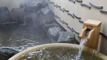 Un baño de aguas termales con agua fluyendo desde un caño de madera hacia una pileta, rodeado de rocas y un muro con detalles curvos.