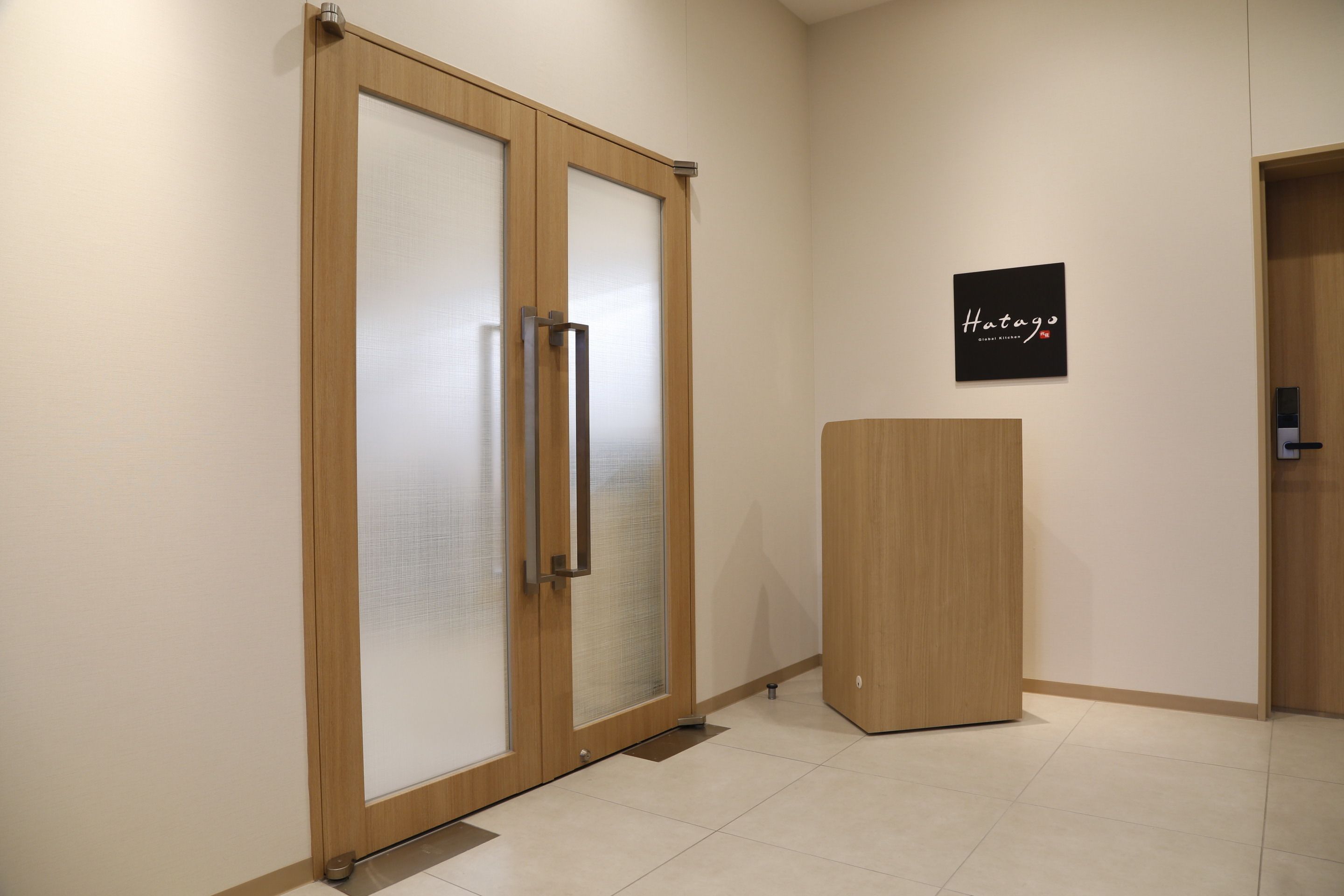 A hallway with a wooden door with frosted glass panels, a small wooden podium, and a sign on the wall that reads "Hatago".