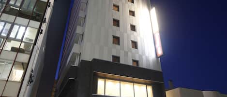 A modern hotel with a prominent entrance, a parking area, and a tall building with illuminated signage in the background.