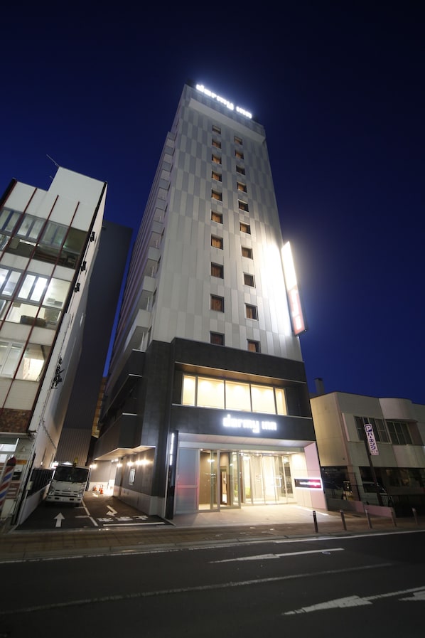 A modern hotel with a prominent entrance, a parking area, and a tall building with illuminated signage in the background.