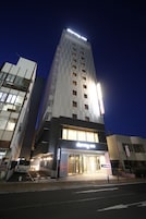 A modern hotel with a prominent entrance, a parking area, and a tall building with illuminated signage in the background.