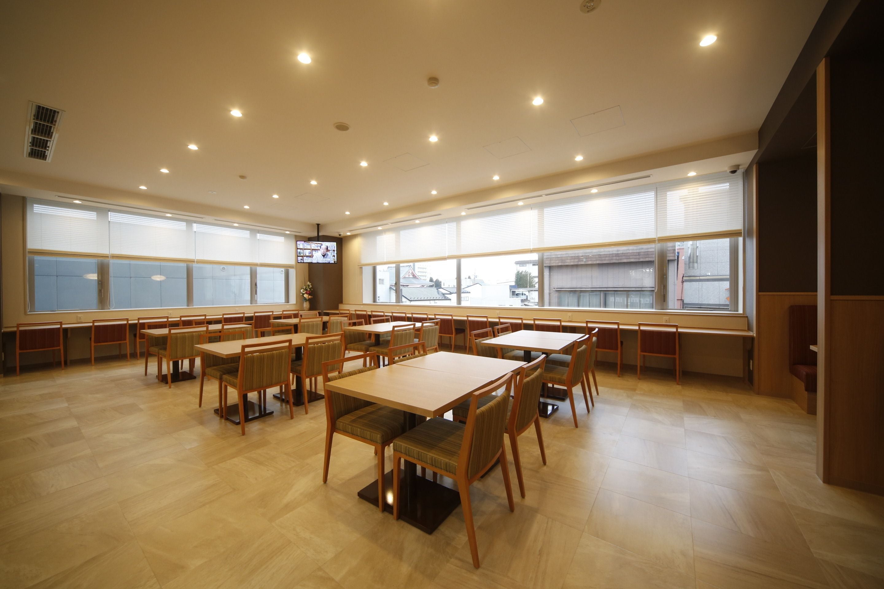 Een ruime eetruimte met meerdere tafels en stoelen, grote ramen met zonwering en inbouwspots in het plafond.