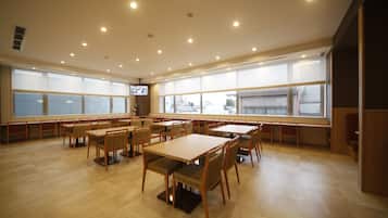 Een ruime eetruimte met meerdere tafels en stoelen, grote ramen met zonwering en inbouwspots in het plafond.