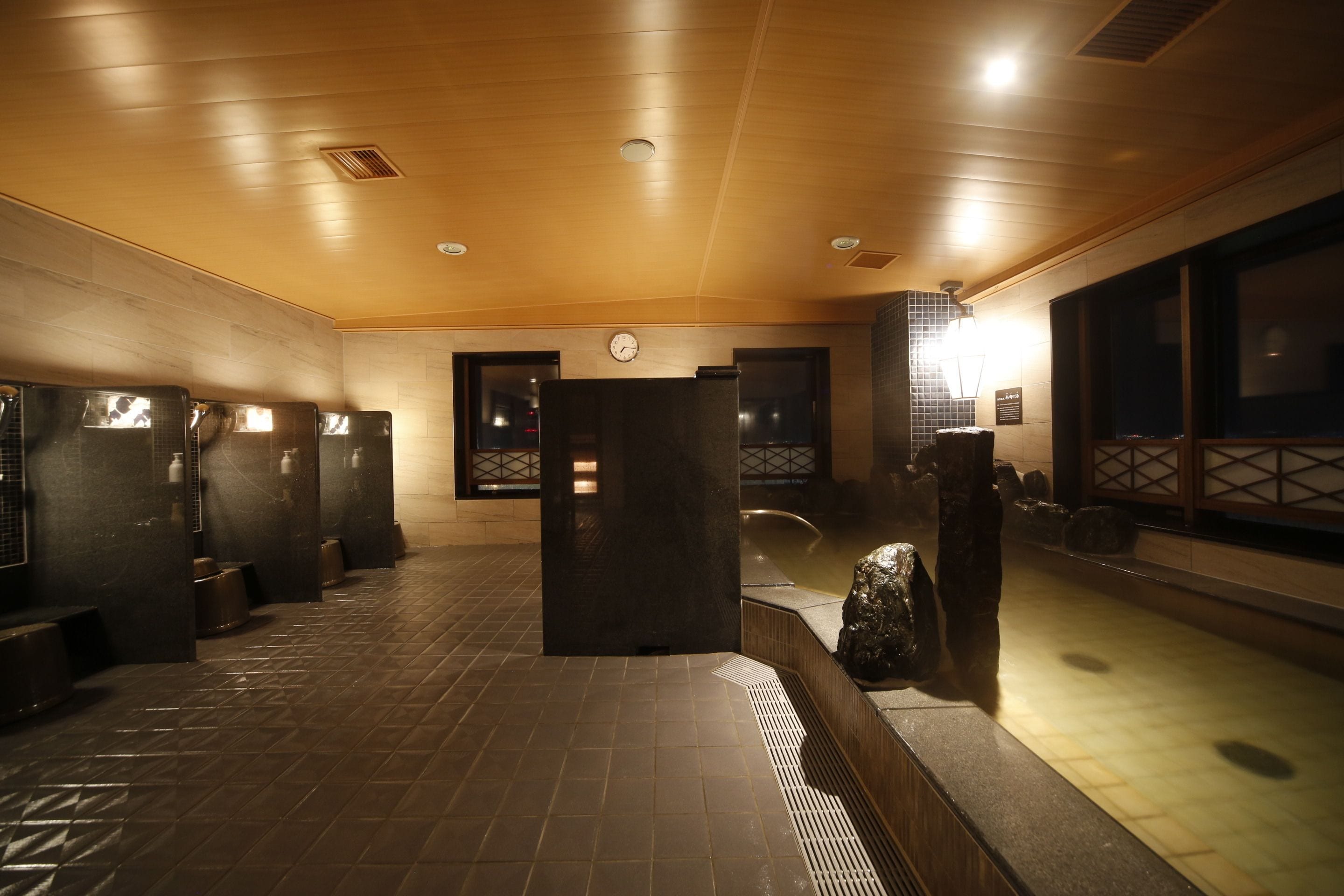 Un baño público con varios cubículos, un reloj en la pared y un ventanal grande a la derecha.