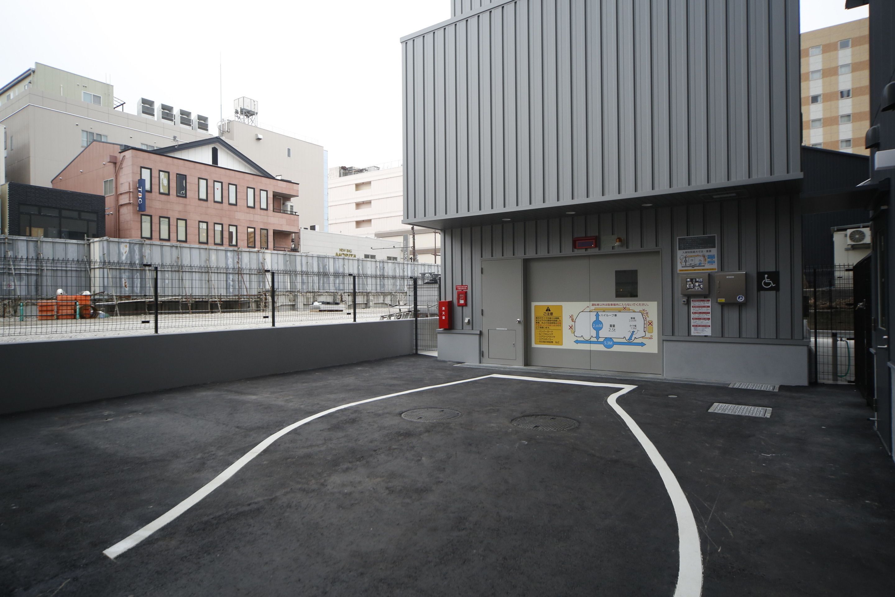 Un espace de stationnement extérieur avec une entrée de bâtiment, une zone délimitée par une clôture et un panneau d’affichage.