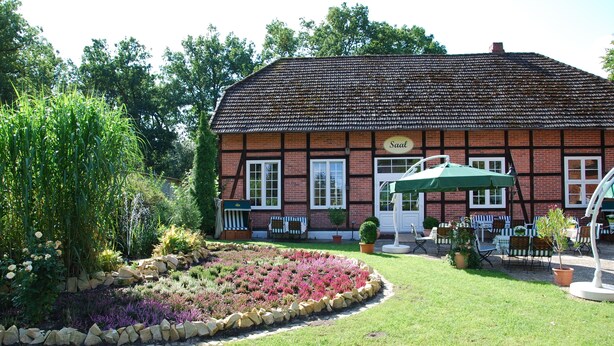 A traditional building with a sign reading "Saal". It features a well-maintained garden with a variety of plants and a stone border. There is a seating area with tables and chairs outside. - Misselhorner Hof (Südheide)
