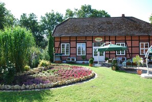 A traditional building with a sign reading "Saal". It features a well-maintained garden with a variety of plants and a stone border. There is a seating area with tables and chairs outside. - Misselhorner Hof (Südheide)