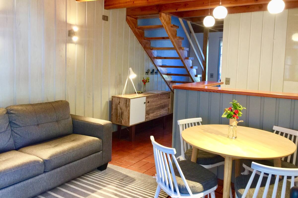 A cozy living room with a grey sofa, a wooden dining table with white chairs, a staircase, and a wooden ceiling.