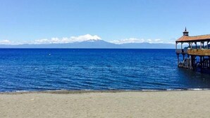A beach with a wooden pier extending into the ocean, a clear sky, and a distant mountain range. - Hotel Frutillar (Frutillar)