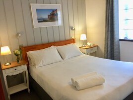 A neatly made bed with white linens, two bedside tables with lamps, a framed picture on the wall, and a window with curtains.