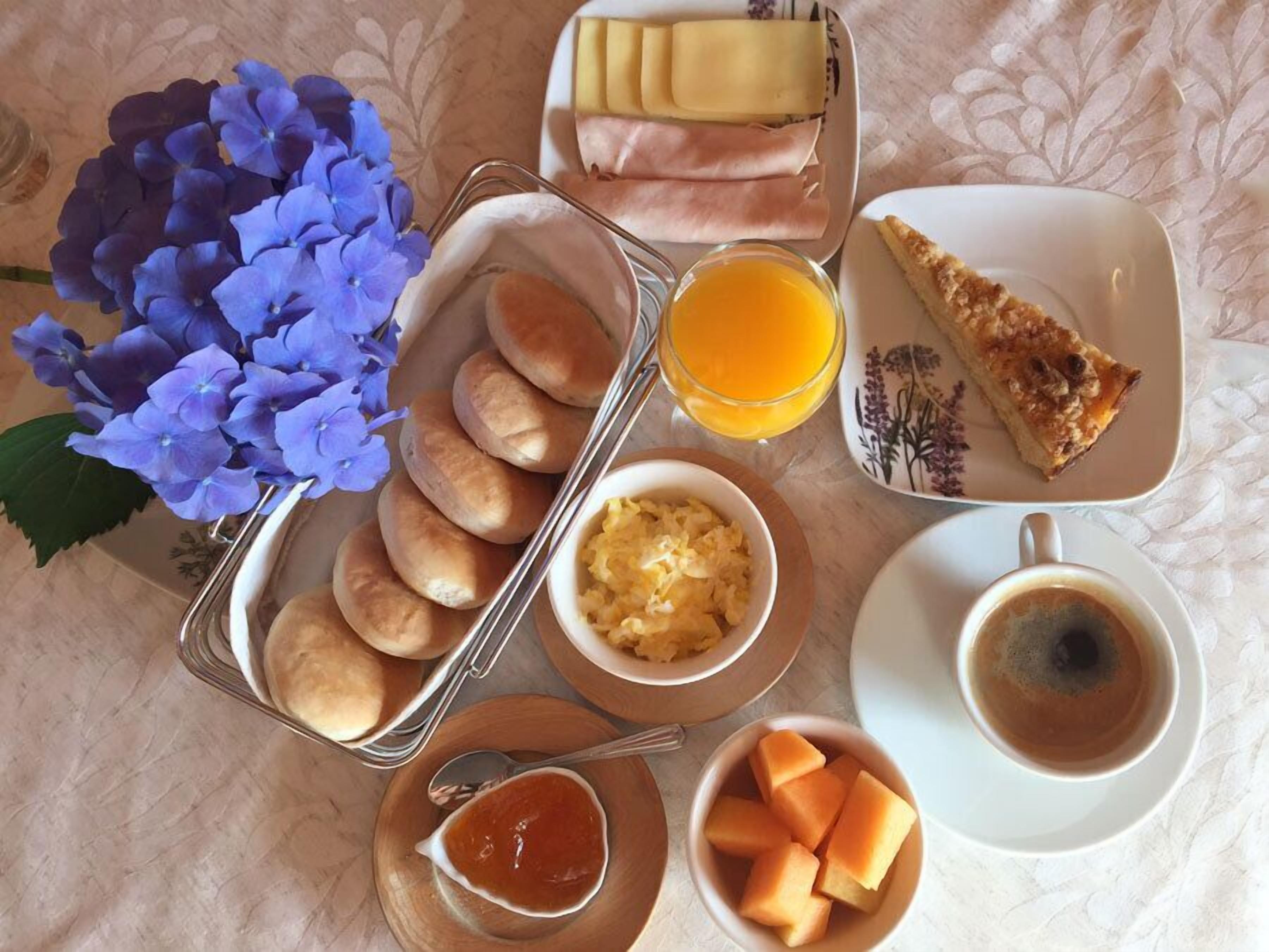 A breakfast setting with bread rolls, cheese, ham, a slice of toast, orange juice, a bowl of scrambled eggs, a bowl of jam, a bowl of cantaloupe, and a cup of coffee.