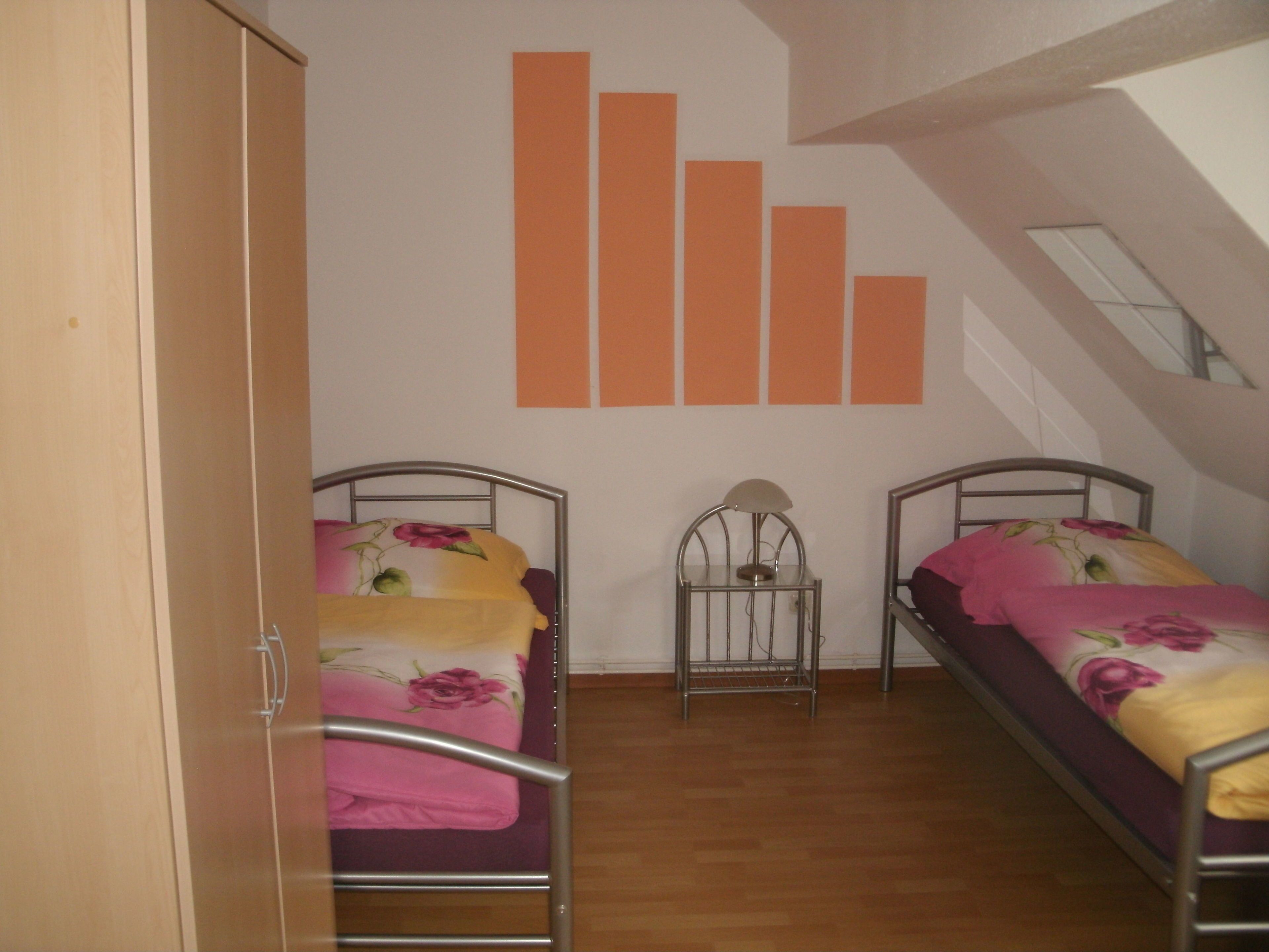 A room with two single beds, a wardrobe, and a wall with orange rectangular decorations.