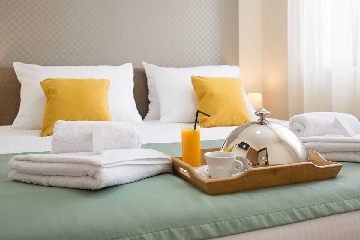 A neatly made bed with white and yellow pillows, a tray with a cup, a glass of orange juice, and a white towel.