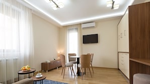 Una sala moderna con un sofá, una pequeña mesa redonda con fruta, una mesa de centro, un área de comedor con sillas, un televisor montado en la pared y un armario de madera.