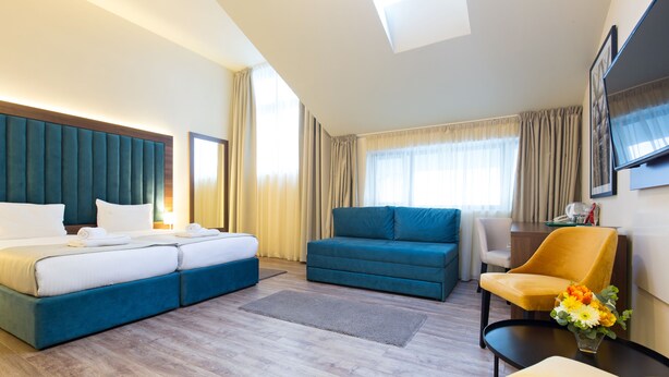 Quarto de hotel com cama, sofá, poltrona, televisão e uma mesinha com um vaso de flores.