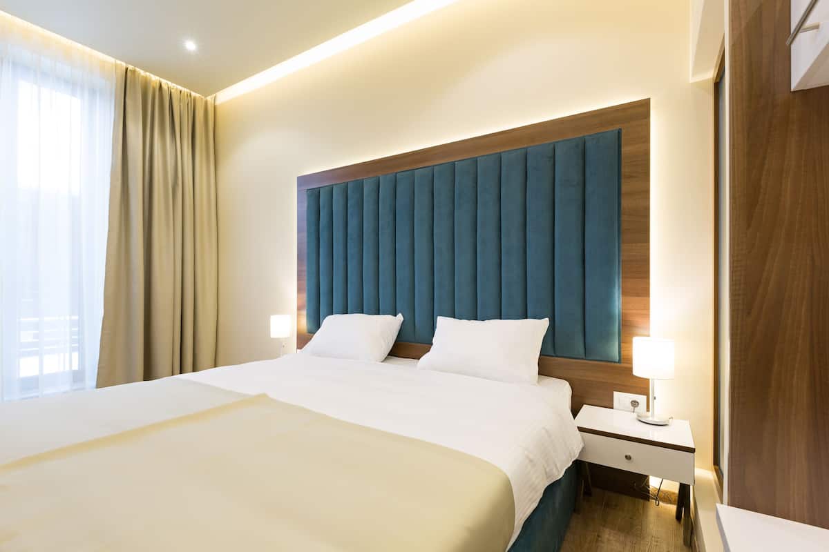 A hotel room with a large bed, a wooden headboard, a nightstand with a lamp, and a window with curtains.